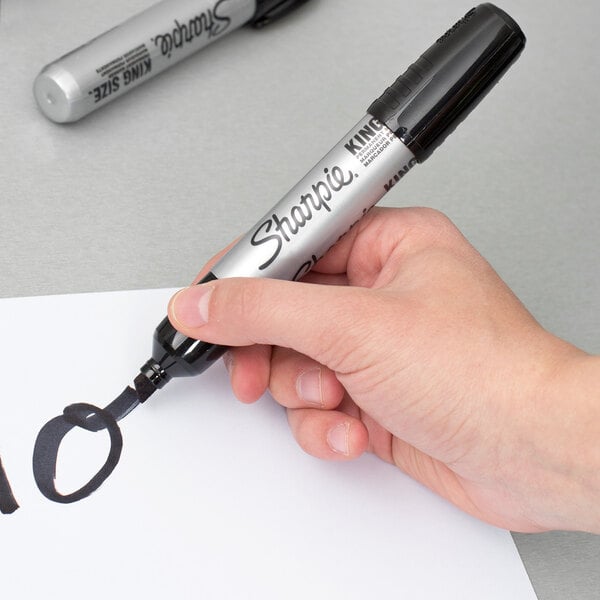 A large black chisel tip Sharpie King Size permanent marker being used to write on white paper.