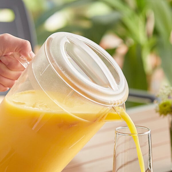 A person pouring orange juice into a Cambro glass pitcher with a clear plastic lid.