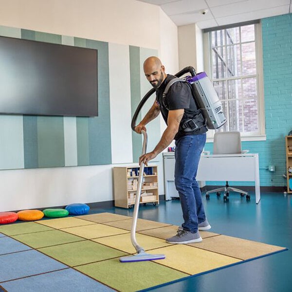A person using a ProTeam GoFit 6 cordless backpack vacuum with a multi-surface wand kit to clean a classroom carpet.