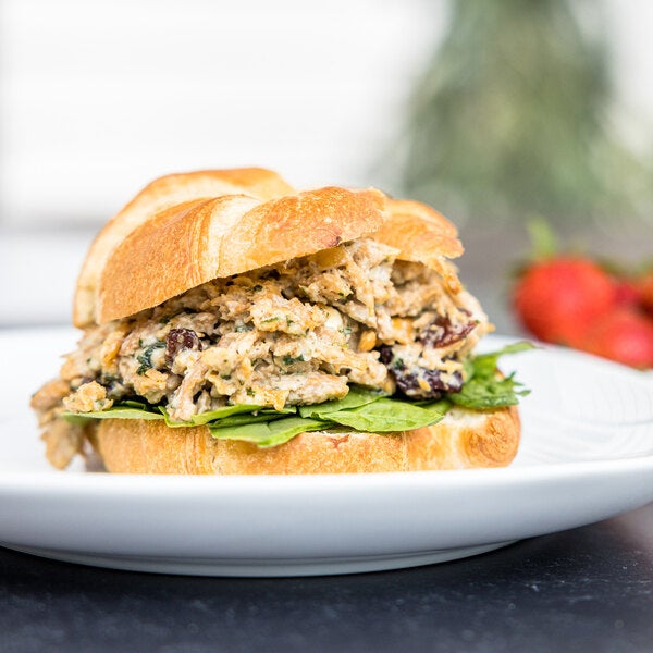 A sandwich made with plant-based vegan wood-smoked shredded chicken and leafy greens on a croissant roll, served on a white plate.