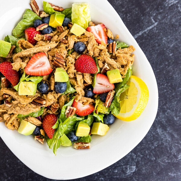 A bowl of salad featuring Barvecue plant-based vegan wood-smoked naked shredded chicken, mixed greens, strawberries, blueberries, avocado, pecans, and a lemon wedge.