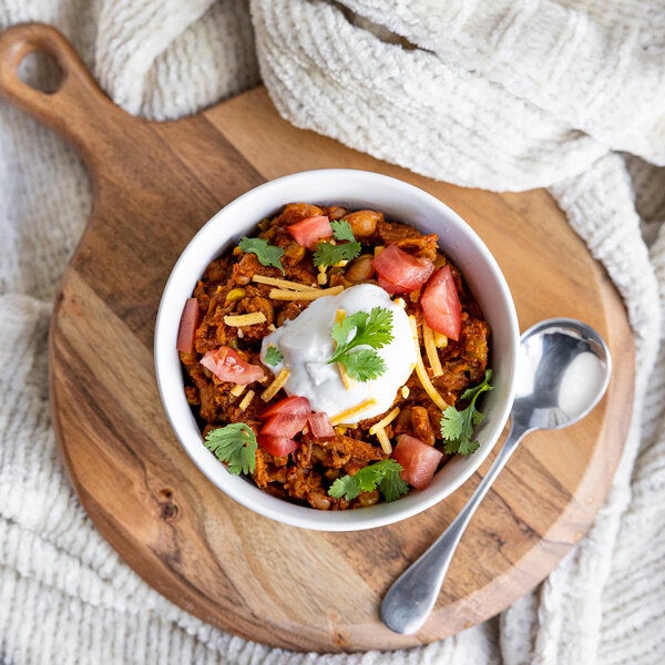 A bowl of plant-based vegan barbecue pulled pork topped with diced tomatoes, shredded cheese, cilantro, and a dollop of cream, served on a wooden board with a spoon.