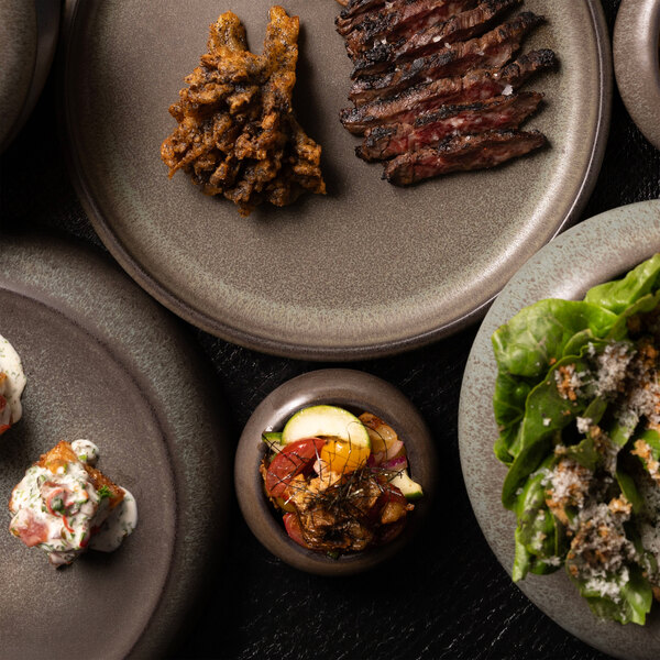 A small round stoneware ramekin filled with a colorful vegetable medley, surrounded by various plated dishes on a dark surface.