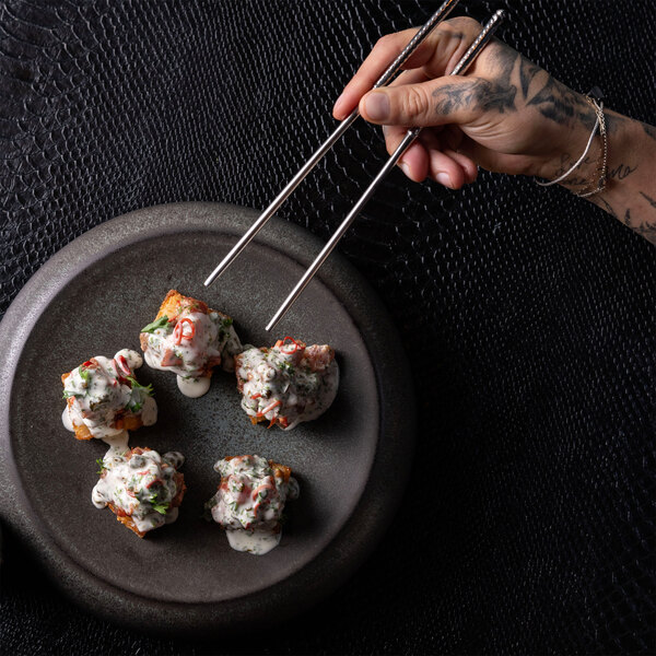 A hand holding chopsticks is picking up one of five pieces of food topped with a creamy sauce and garnished with herbs and sliced chili, served on a round, dark stoneware plate.
