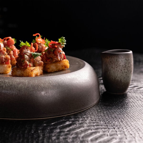 A small, round, green and brown stoneware creamer sits next to a dark, modern plate with gourmet appetizers on a textured black surface.