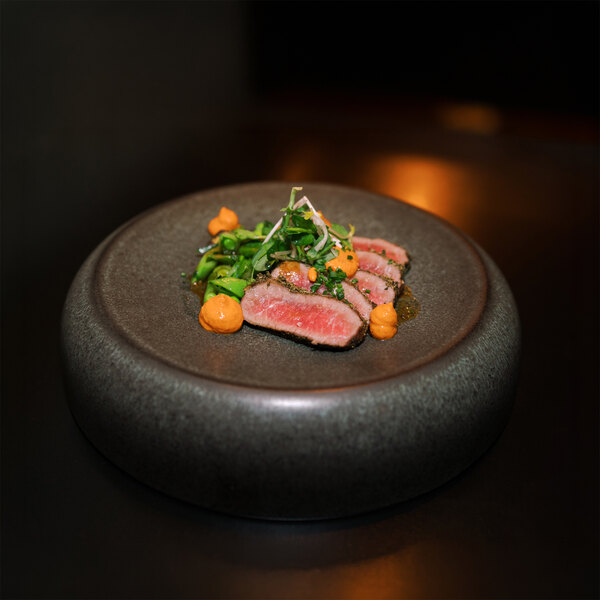 A round, elevated dark stoneware plate displaying a small, artfully arranged portion of sliced meat, green vegetables, and orange sauce dollops.
