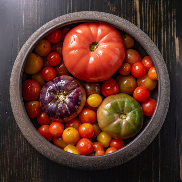 A round stoneware bowl filled with a variety of colorful tomatoes, including red, yellow, green, and purple ones, placed on a dark wooden surface.