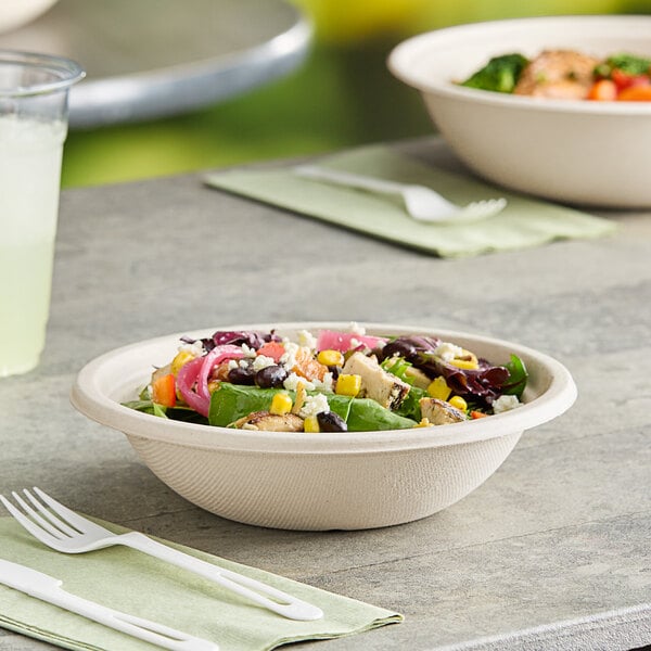 A compostable fiber take-out bowl filled with salad, placed on a table with utensils and a drink nearby.