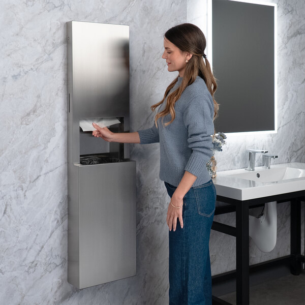 A stainless steel surface-mounted paper towel dispenser and waste receptacle with a satin finish, installed in a restroom.
