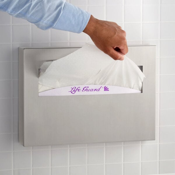 A stainless steel surface-mounted seat cover dispenser with a satin finish, installed on a tiled wall.