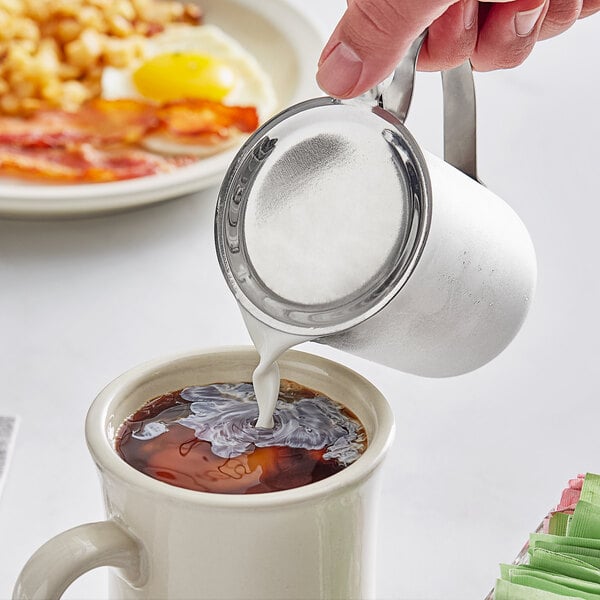 A stainless steel creamer with a closed handle being used to pour cream into a cup of coffee.