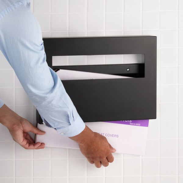 A matte black surface-mounted seat cover dispenser being used to dispense a paper seat cover.
