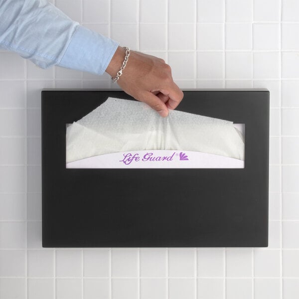 A matte black surface-mounted seat cover dispenser being used to pull out a paper seat cover.