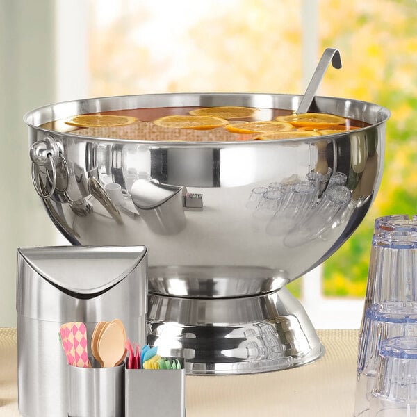 A large stainless steel punch bowl with handles and a mirror finish, filled with punch and garnished with citrus slices.