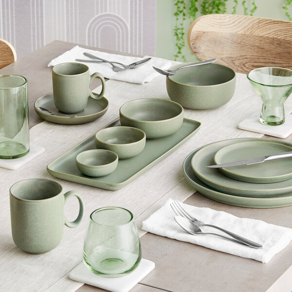 A dining table set with sage green matte finish porcelain dishes, bowls, mugs, and green-tinted glassware.