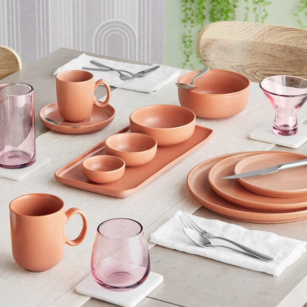 A table set with terra cotta-colored porcelain dinnerware, including bowls, plates, mugs, and pink-tinted glassware.