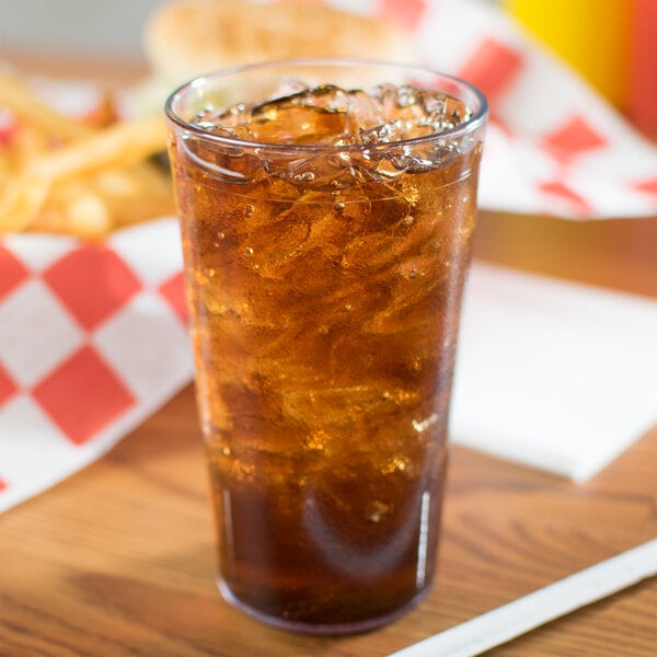 A Carlisle clear plastic tumbler filled with brown liquid and ice on a table with a sandwich and fries.