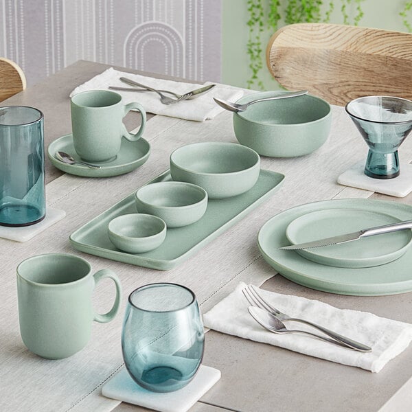 A neatly set dining table featuring matte blue-green porcelain dishes, bowls, mugs, and a variety of blue-tinted glassware.