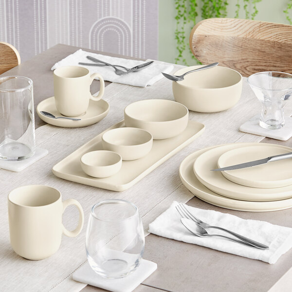A dining table set with cream-colored porcelain dishes, bowls, mugs, and various glassware, arranged neatly with silverware on white napkins.