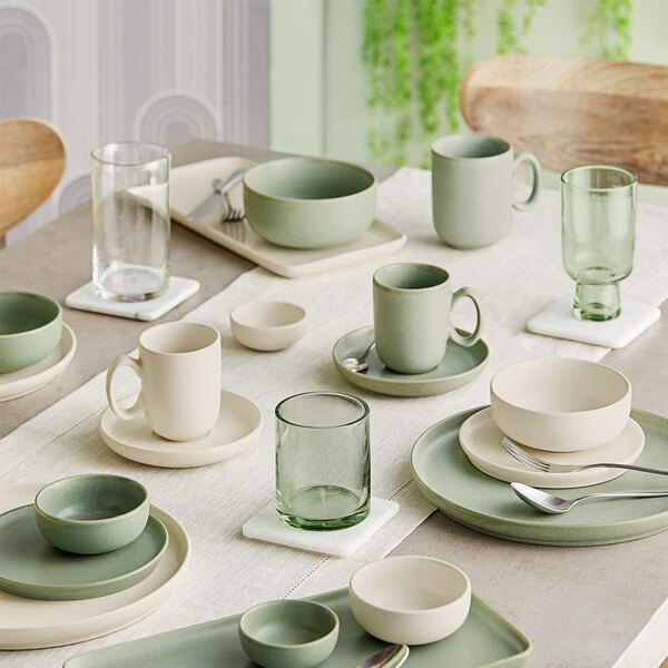 A neatly arranged dining table set with green and white matte porcelain plates, bowls, mugs, and clear glassware on a light tablecloth.