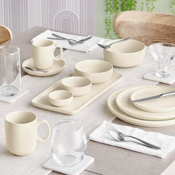 A neatly set dining table featuring cream-colored porcelain dishes, bowls, mugs, and various glassware with silverware placed on white napkins.