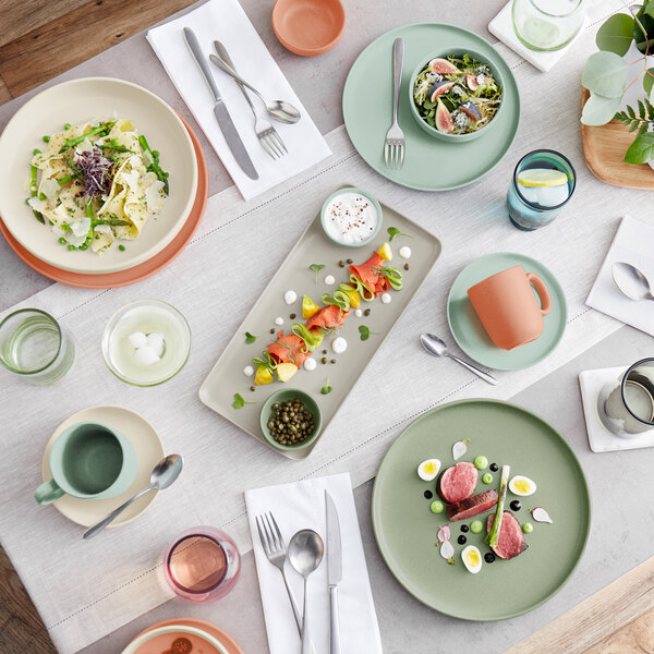 A table set with assorted pastel-colored plates, bowls, cups, and various dishes, including salads, meat, and skewers, arranged in a modern and elegant style.