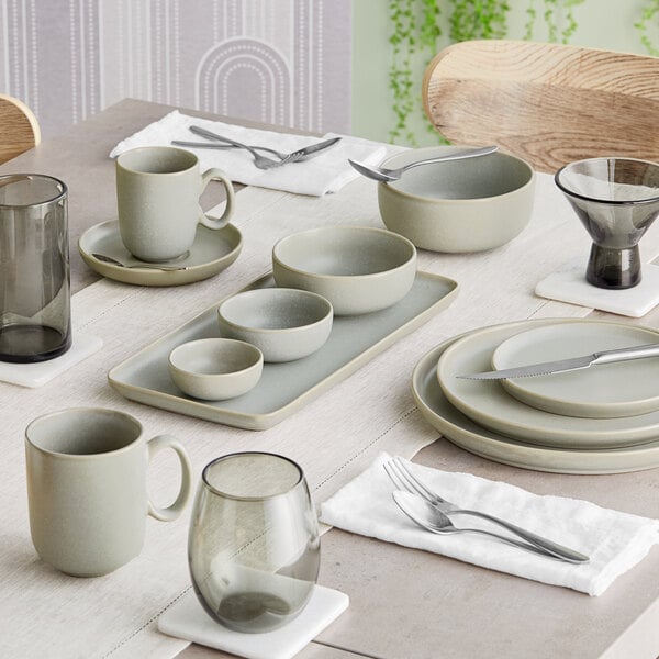 A set dining table featuring ash gray matte finish porcelain mugs, bowls, plates, and glassware arranged neatly with cutlery and napkins.