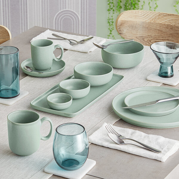 A neatly set dining table featuring matte blue-green porcelain dishes, bowls, mugs, and a variety of blue-tinted glassware.