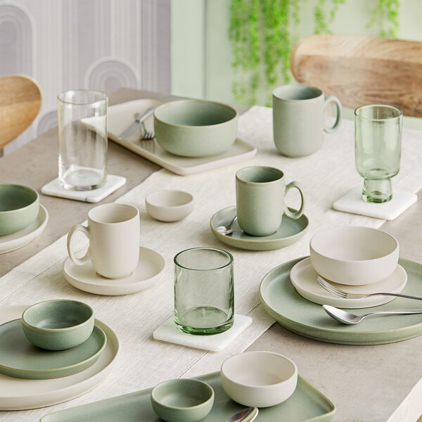 A table set with sage green and cream-colored porcelain cups, bowls, plates, and green-tinted glassware arranged neatly on a light tablecloth.