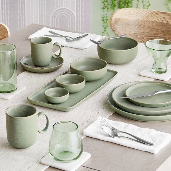A table set with sage green matte finish porcelain dishes, cups, and bowls, accompanied by green glassware and silverware on white napkins.