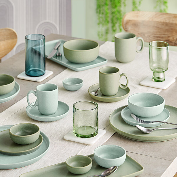 A dining table set with various pastel-colored ceramic dishes, bowls, mugs, and green-tinted glassware arranged neatly on a light tablecloth.