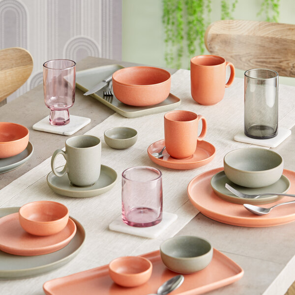A table set with various pastel-colored dishes, bowls, mugs, and glasses in shades of orange, gray, and pink, arranged neatly on a light tablecloth.