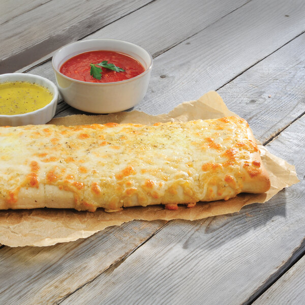 A rectangular slab of cheesy breadsticks served on parchment paper with two dipping sauces, one red and one yellow.