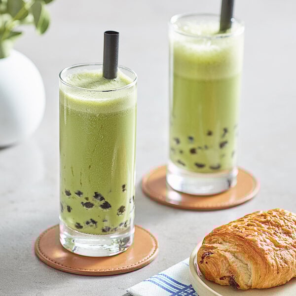 A pair of tall glasses filled with green matcha bubble tea, each with a black straw, placed on coasters next to a chocolate croissant.