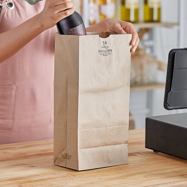 A woman opening a bottle of wine in a brown paper bag on a table.