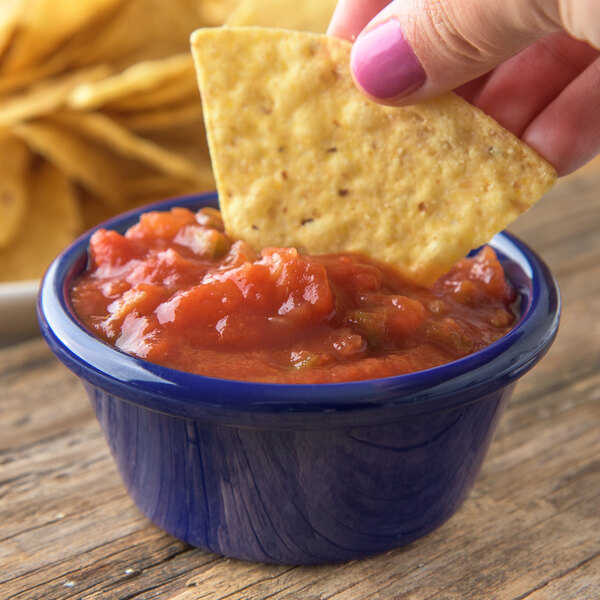 A person holding a blue chip and dipping it into a blue Carlisle ramekin filled with salsa.