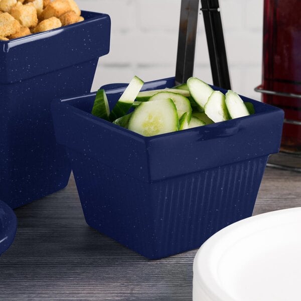 A blue Tablecraft square condiment bowl with cucumbers and crackers on a counter.