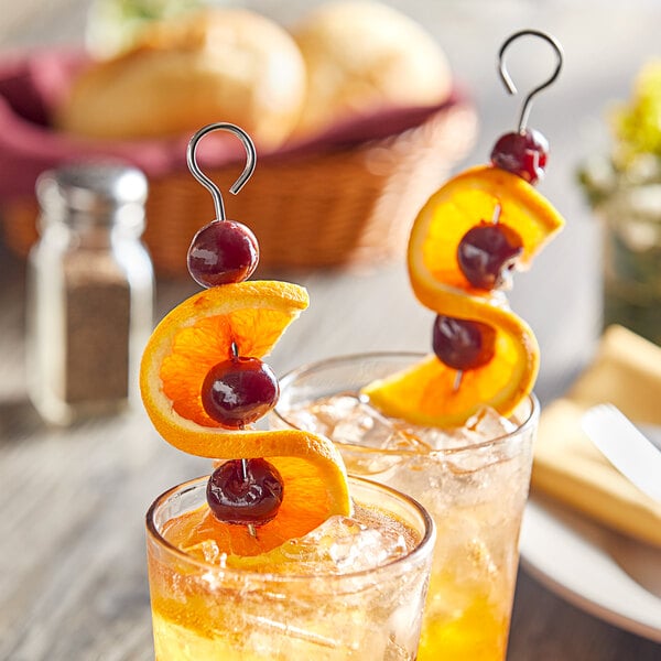 A stainless steel flat skewer garnished with orange slices and cherries, placed in a glass of iced beverage.