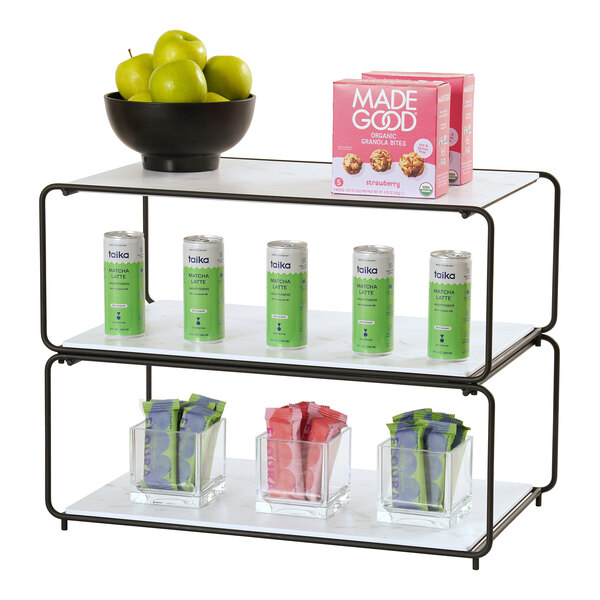 A three-tiered modular display rack holding a bowl of green apples, boxes of granola bites, cans of matcha latte, and clear containers with assorted snack packs.