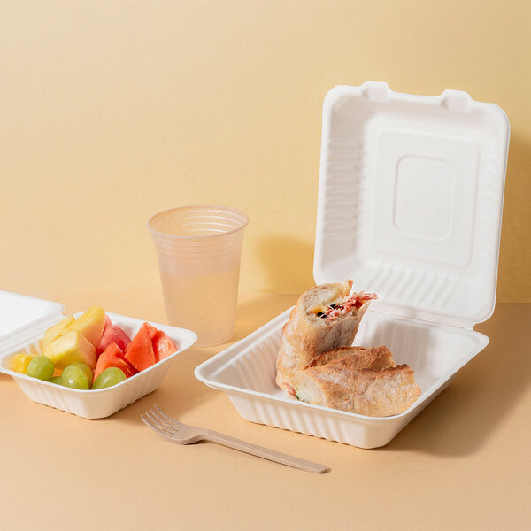 A white compostable sugarcane fiber clamshell take-out container holding a sandwich, shown alongside a fruit container, plastic cup, and wooden fork.