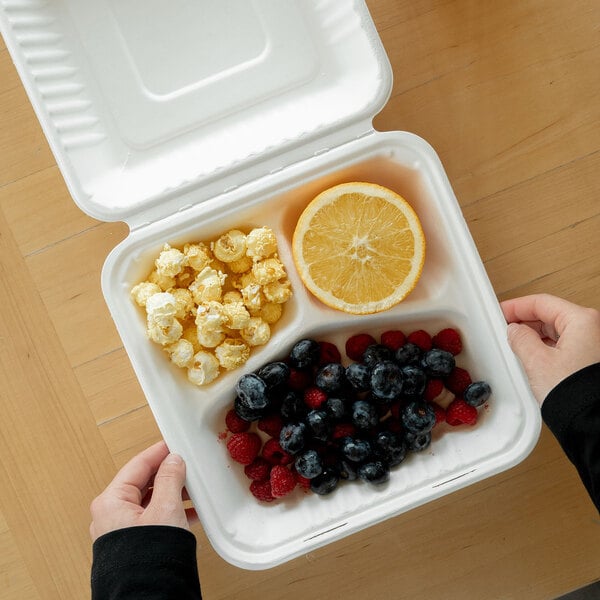 A white, three-compartment compostable clamshell take-out container made from sugarcane fiber, shown holding popcorn, a halved orange, and mixed berries.