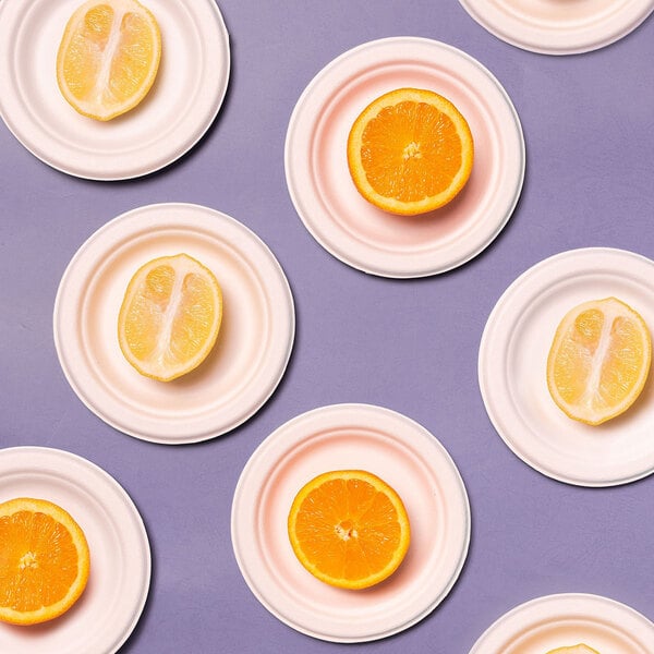 A set of small round white sugarcane fiber plates, each holding either a halved orange or lemon, arranged on a purple surface.