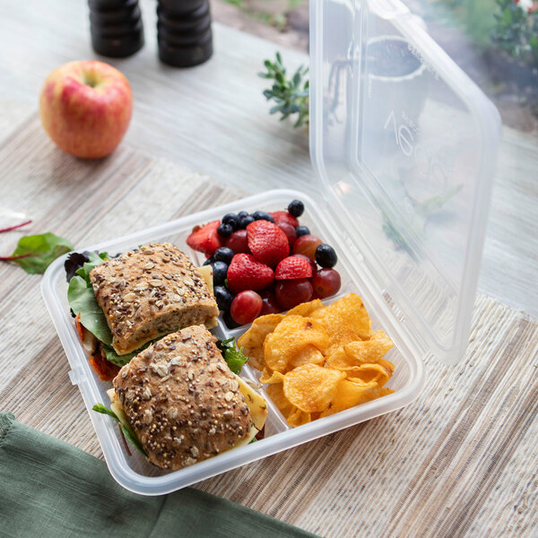 A clear, three-compartment hinged reusable take-out container holding two sandwiches, mixed berries, and chips.