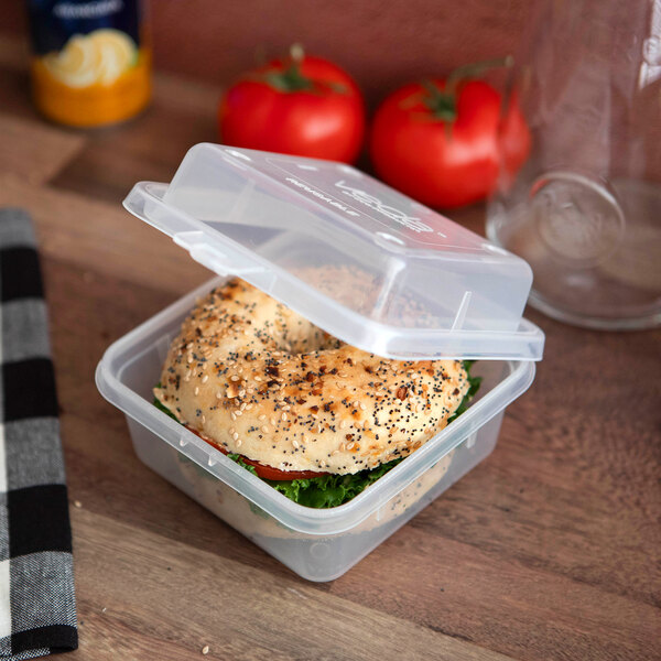 A clear, hinged, reusable take-out container holding a sandwich, shown on a wooden surface.