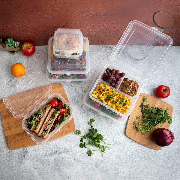 A clear, three-compartment hinged reusable take-out container filled with food, shown alongside other similar containers and fresh ingredients.