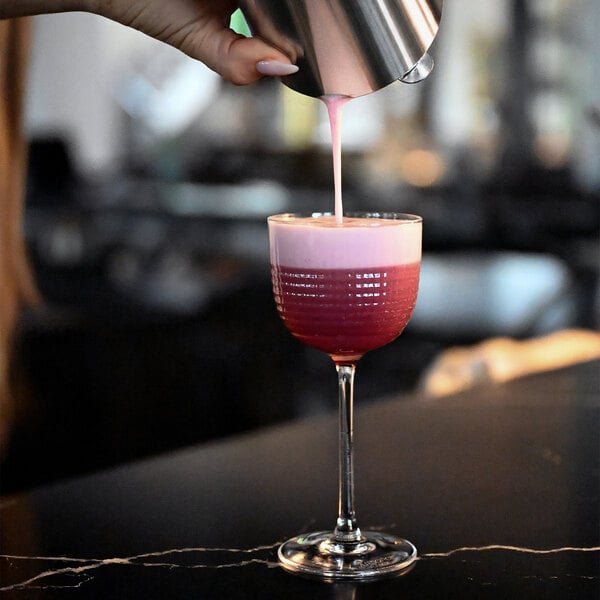 A hand is pouring a pink foamy liquid from a metal shaker into a clear, textured stemmed glass on a dark surface.
