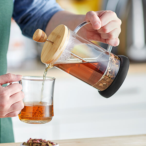 A person is pouring tea or coffee from a glass French press with a wooden lid into a clear glass cup.