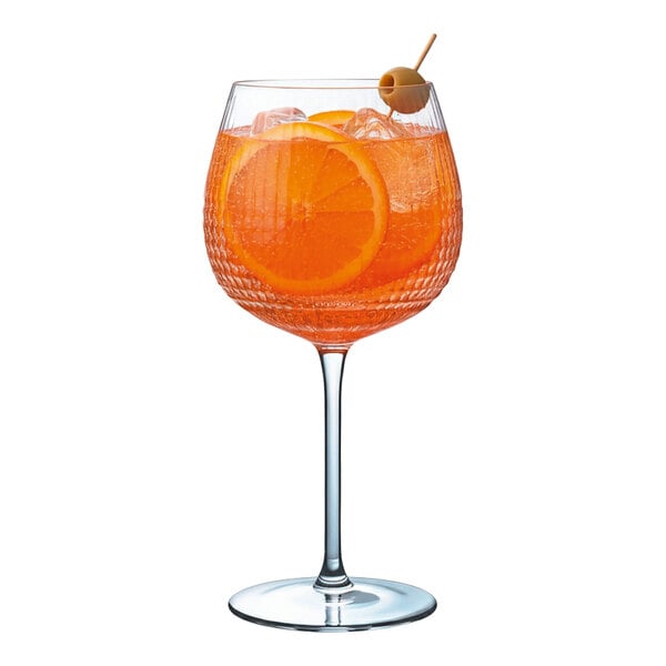 A textured 20 oz. gin and tonic glass filled with an orange cocktail, ice, orange slices, and garnished with an olive on a pick.