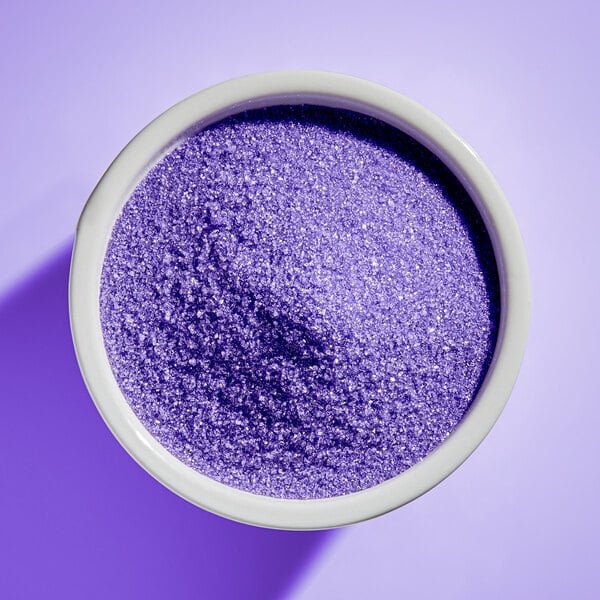 A white bowl filled with fine, sparkling lavender-colored sugar granules on a lavender background.
