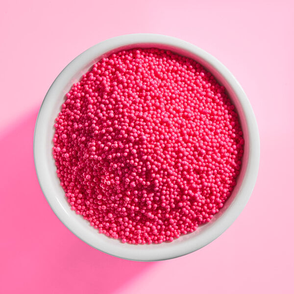 A white bowl filled with small, round, bright pink sprinkles on a pink background.